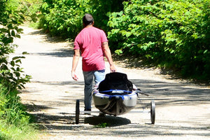 Man in red shirt pulling a gray Sea Eagle kayak on a white EZ Cart with black wheels down a dirt path.