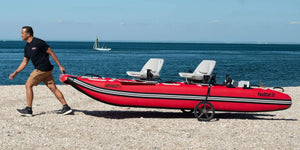 Man pulling a red Sea Eagle FastCat12 inflatable catamaran with two white seats on a pebbly beach.