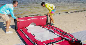 Man and boy assembling a red Sea Eagle FastCat14 inflatable catamaran on a sandy beach.