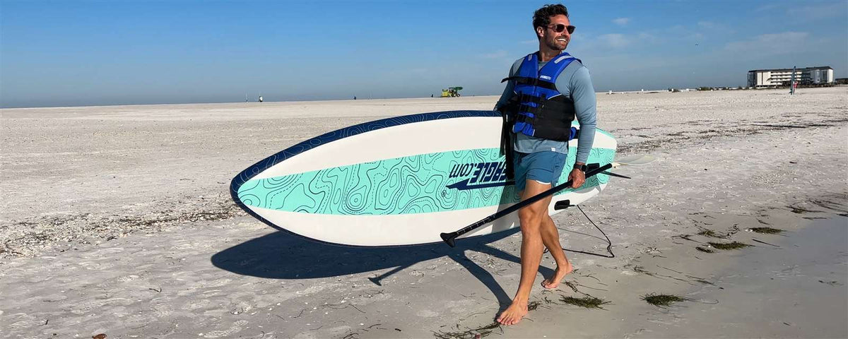 Man in a life vest carrying a white, blue, and teal Sea Eagle LongBoard 11 inflatable paddleboard and paddle on a sandy beach.