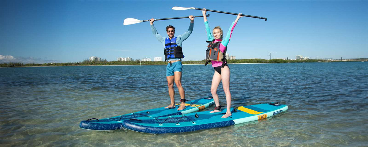 Two people stand on blue and teal Sea Eagle LongBoard 11 inflatable paddleboards, holding paddles overhead.
