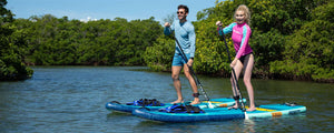 Two people paddle blue Sea Eagle LongBoard 11 inflatable SUPs on calm water with green trees.