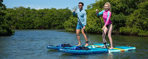 Two people paddle blue Sea Eagle LongBoard 11 inflatable SUPs on calm water with green trees.