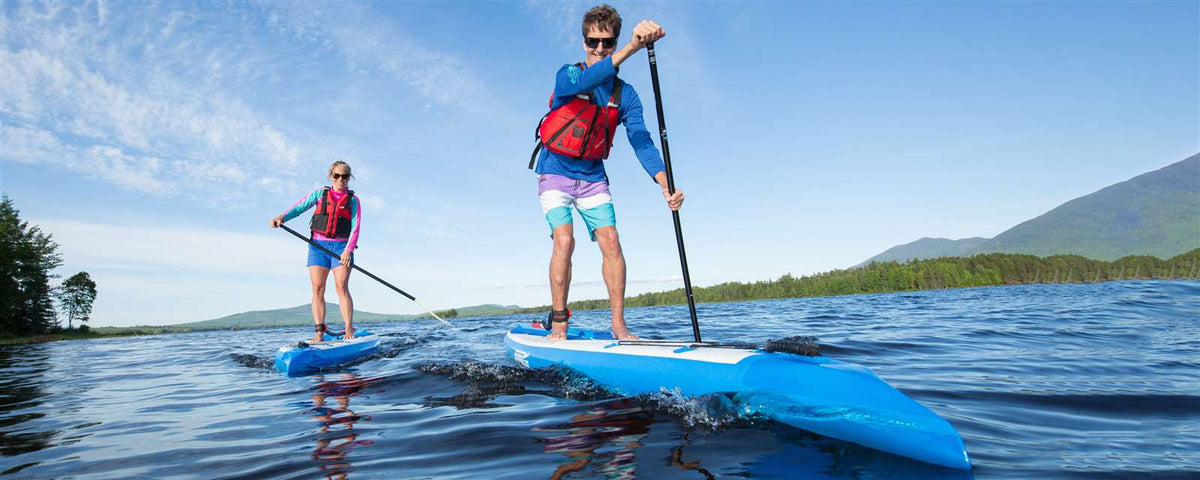 Two people paddle blue Sea Eagle NeedleNose 14 inflatable SUPs on a lake with mountains.