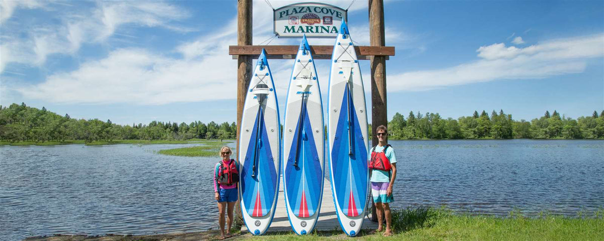 Three blue and white Sea Eagle NeedleNose™ inflatable paddleboards with paddles lean against a wooden marina sign.