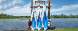 Three blue and white Sea Eagle NeedleNose™ inflatable paddleboards with paddles lean against a wooden marina sign.