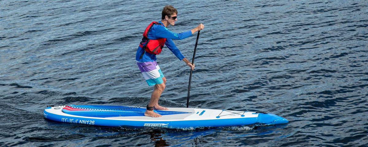 Man paddling a blue and white Sea Eagle NeedleNose 126 inflatable paddleboard on calm water.