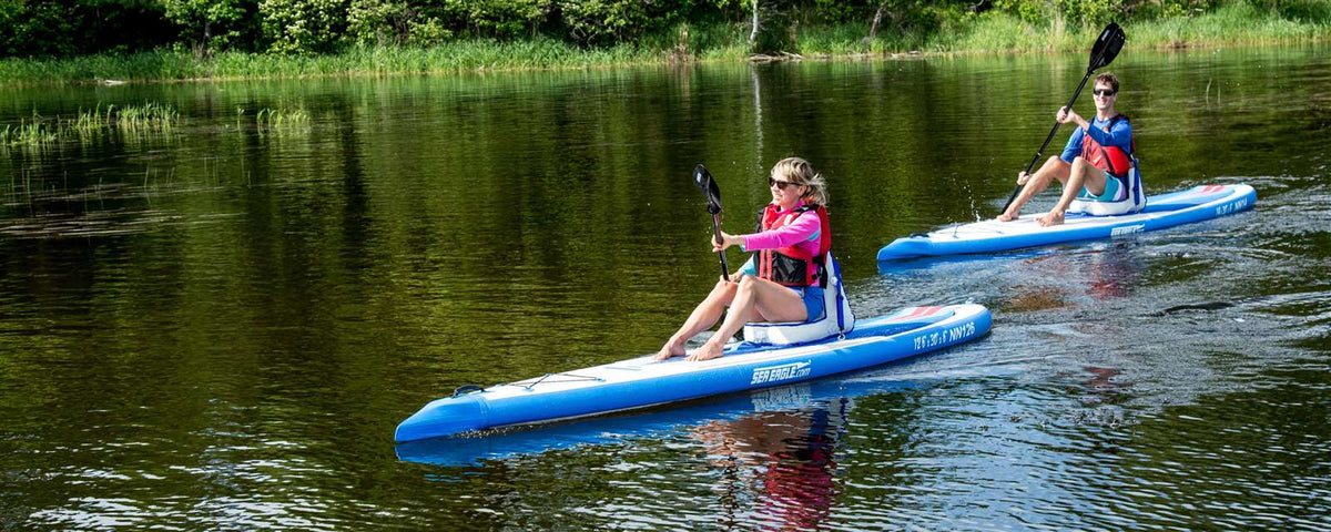 Two people paddling blue and white Sea Eagle NeedleNose 126 inflatable paddleboards on a calm lake.