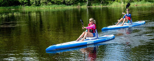 Two people paddling blue and white Sea Eagle NeedleNose 126 inflatable paddleboards on a calm lake.