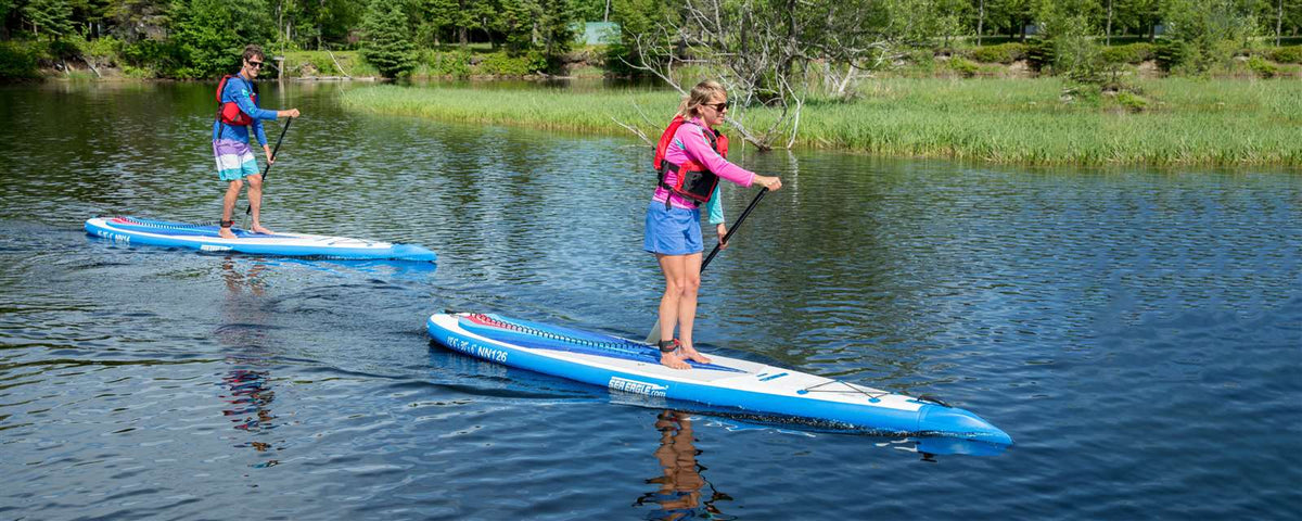 Two people paddle blue and white Sea Eagle NeedleNose 126 inflatable paddleboards on a calm lake.