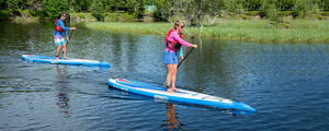 Two people paddle blue and white Sea Eagle NeedleNose 126 inflatable paddleboards on a calm lake.