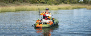 Man in an orange shirt and black vest holding a fish in a green and orange Sea Eagle PackFish7™ inflatable boat.