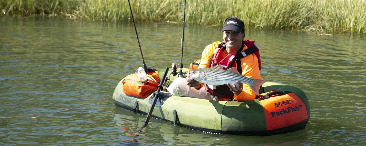 Man in an orange shirt and life vest holding a striped bass in a green Sea Eagle PackFish7™ inflatable fishing boat.