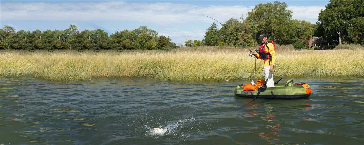 Angler standing in an olive green Sea Eagle PackFish7™ inflatable boat, reeling in a fish in a grassy marsh.