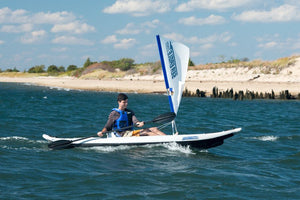 Man sailing a white kayak with a blue and white Sea Eagle QuikSail on open water near a sandy shore.