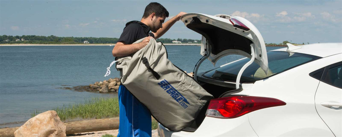 Man loading a compact, rolled-up Sea Eagle inflatable boat in a gray storage bag into a white car trunk by the water.