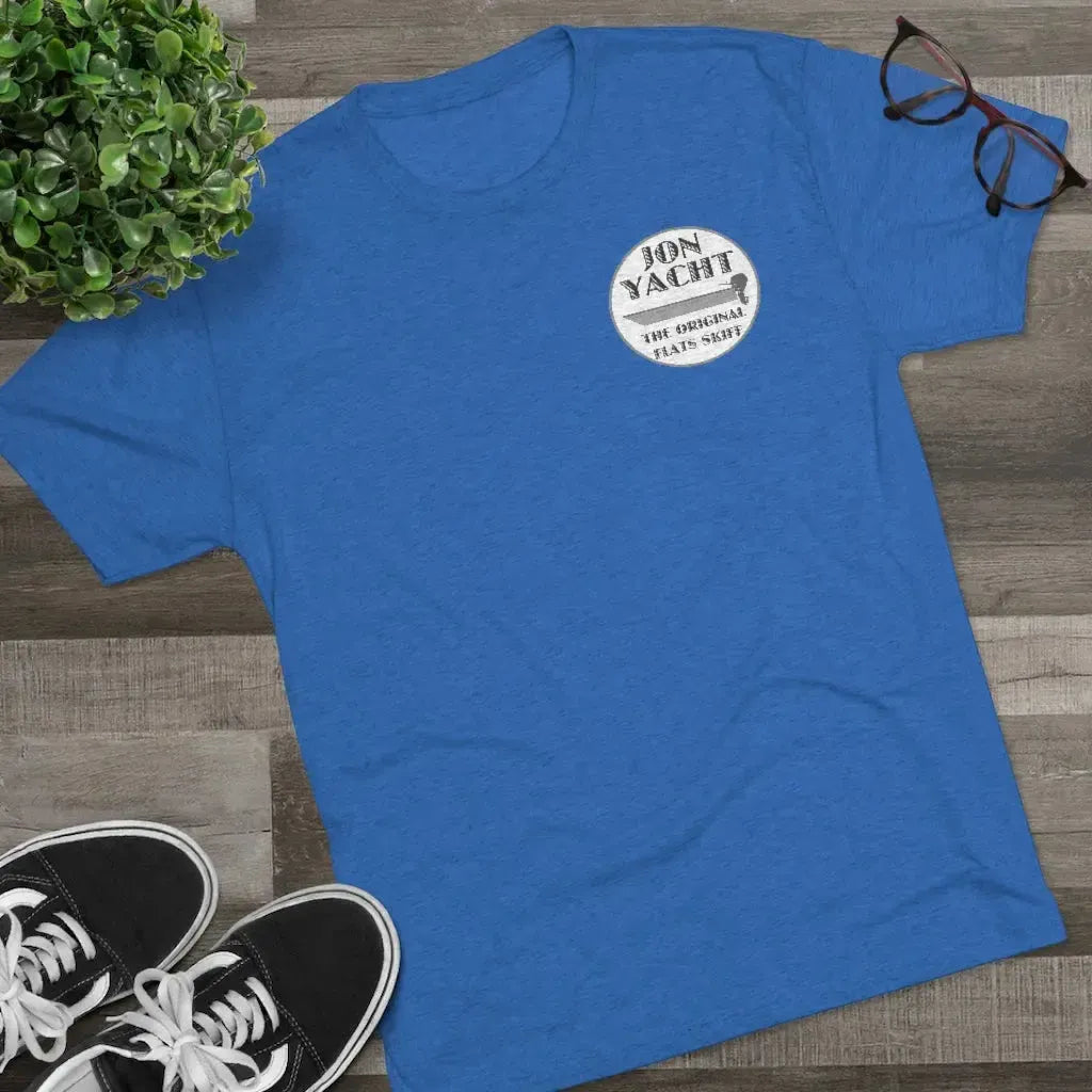 Blue "Jon Yacht The Original Flats Skiff" graphic tee, black sneakers, plant, and glasses on wood.