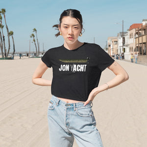 Woman in a black flowy cropped tee with "JON YACHT" text, light wash jeans, on a sunny beach.