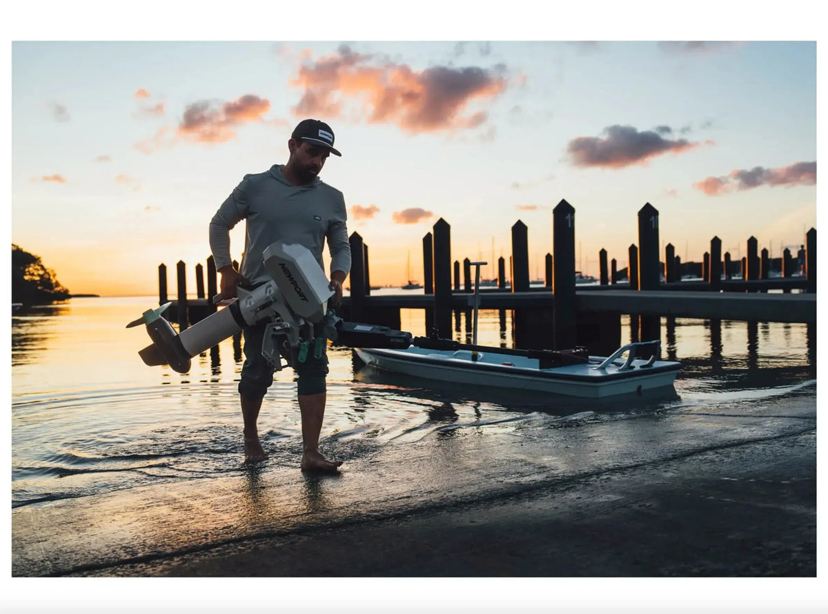 Man carries a gray Newport NT600 6hp electric outboard motor into the water at sunset, next to a small boat.