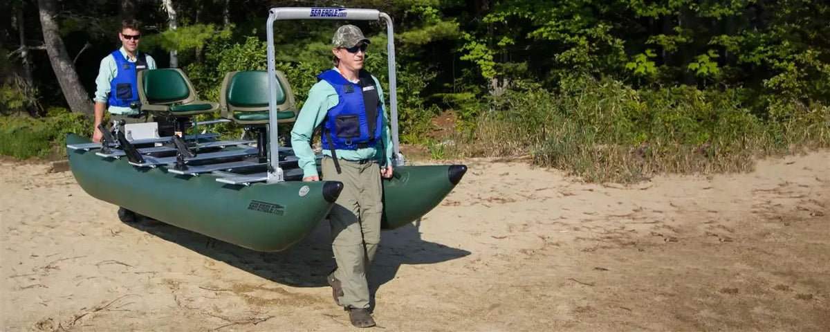 Two men carry a dark hunter green Sea Eagle 375fc FoldCat inflatable fishing boat with two seats on a sandy beach.