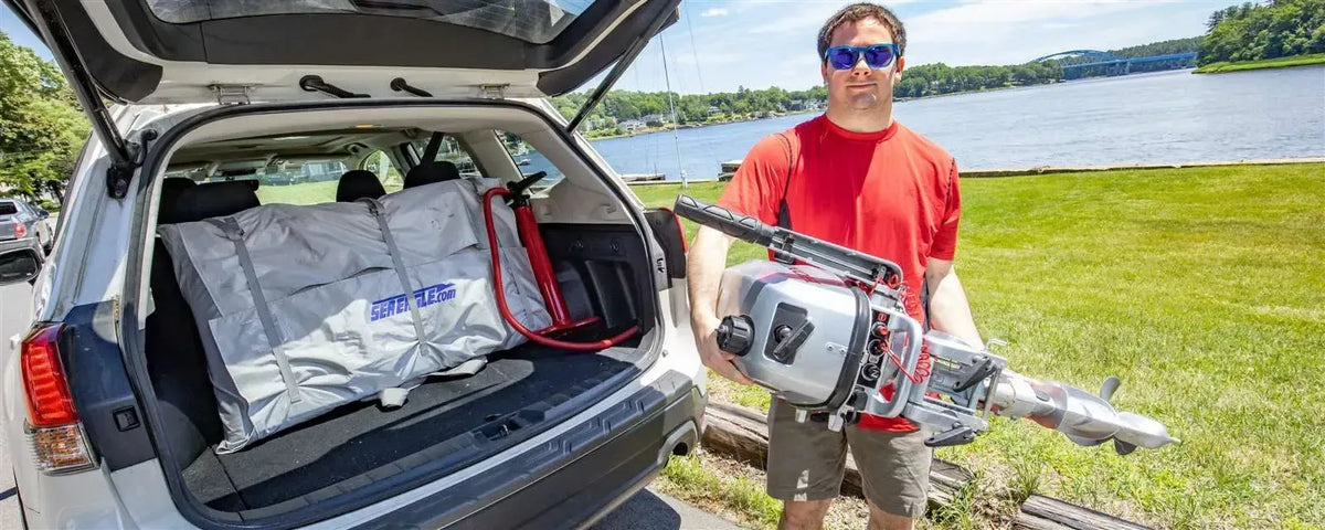Man holding a silver outboard motor next to a white SUV with a packed Sea Eagle inflatable boat.