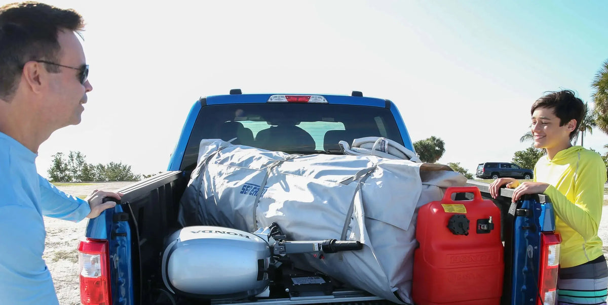 Two men loading a rolled-up gray Sea Eagle FastCat14 inflatable catamaran, silver Honda outboard motor, and red fuel tank into a blue pickup truck bed.
