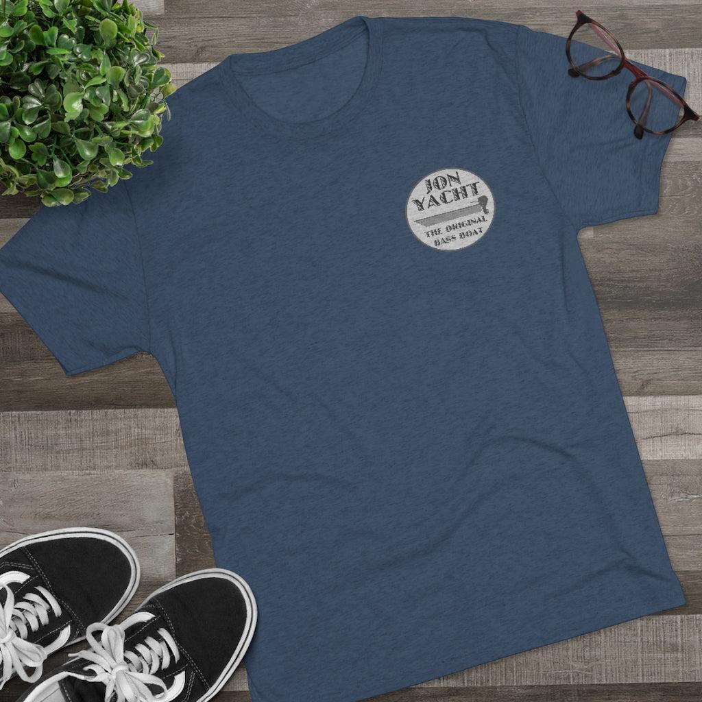 A heathered blue crewneck t-shirt with a circular white graphic on the left chest reading 'JON YACHT'. The shirt is styled in a flat-lay arrangement on a gray wood-plank floor, accompanied by black sneakers, eyeglasses, and a green potted plant.