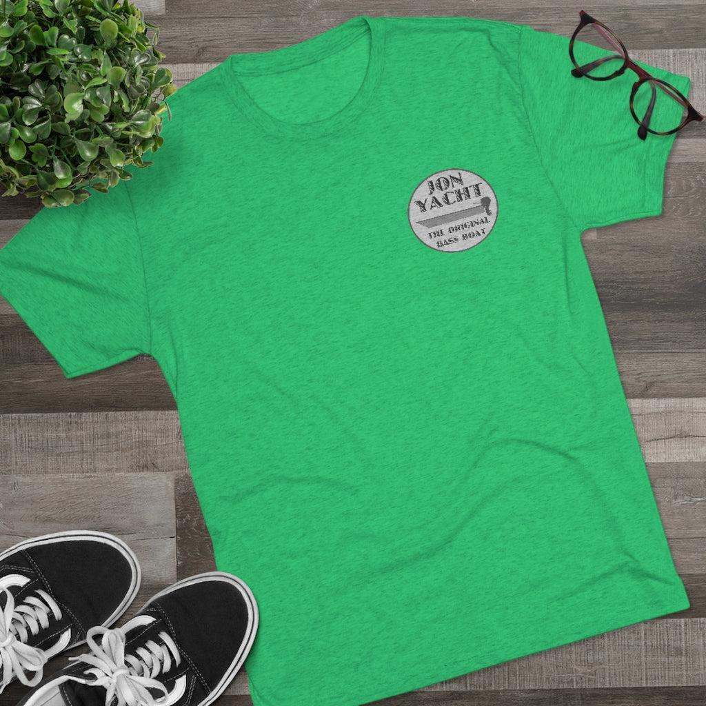 A bright green heathered t-shirt laid flat on a gray wood plank surface, styled with a potted plant, black eyeglasses, and black sneakers. The shirt features a circular graphic on the left chest that reads 'JON YACHT'.