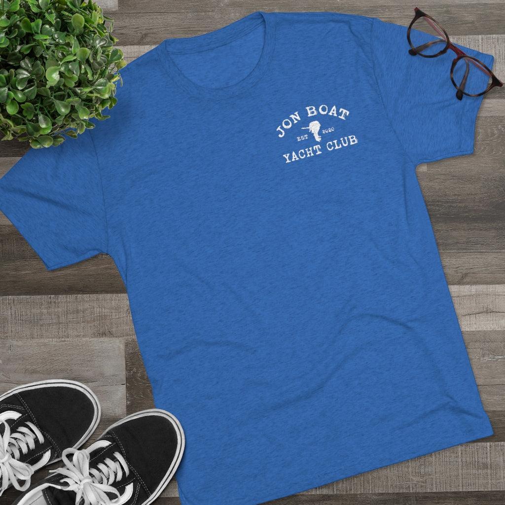 A heather blue short-sleeve t-shirt with a white graphic reading 'JON BOAT YACHT CLUB' on the left chest, styled in a flat lay on a wood floor with a green plant, eyeglasses, and black sneakers.