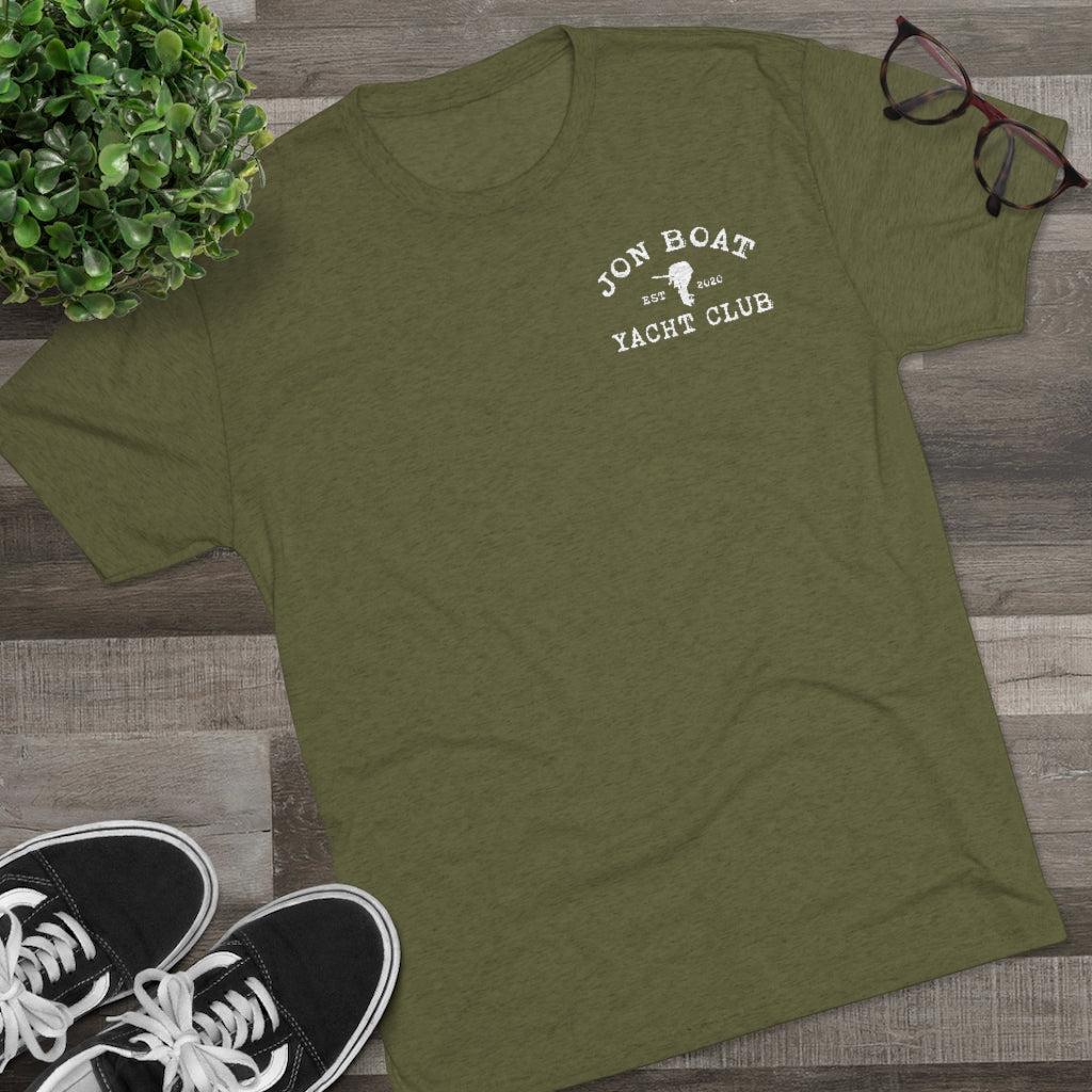 An olive green heather t-shirt with a white graphic reading 'JON BOAT YACHT CLUB EST. 2024' on the left chest, styled in a flat lay on a gray wood floor with black sneakers, eyeglasses, and a green plant.