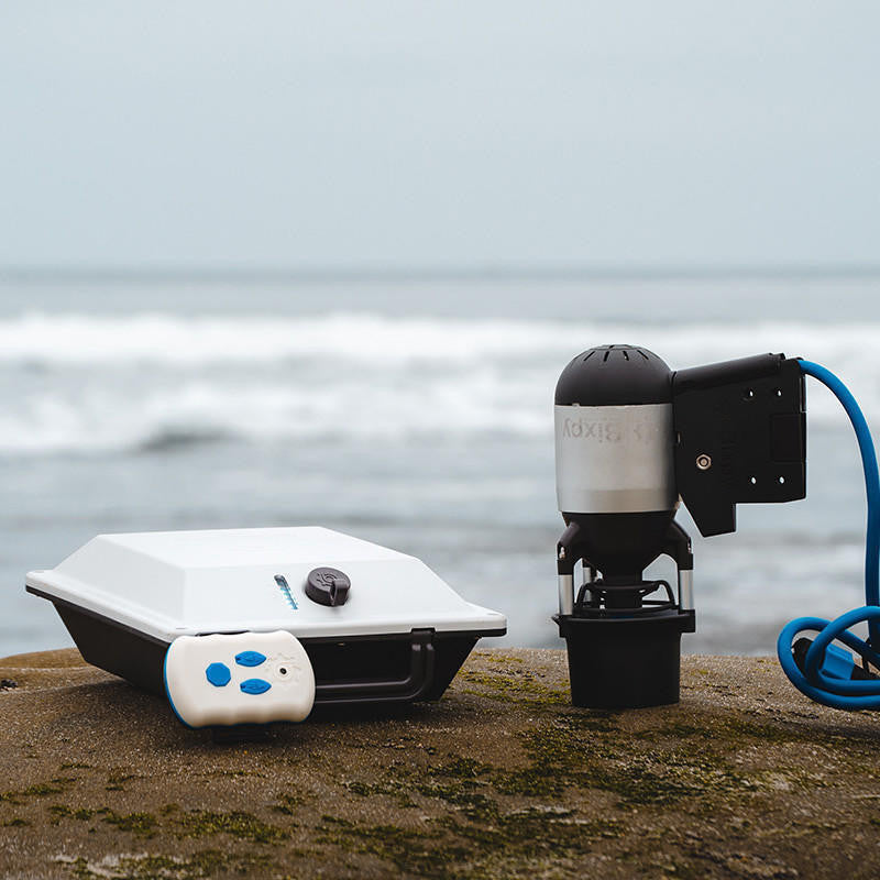 Two devices, one white and one black with a hose, on a rocky surface with water in the background.