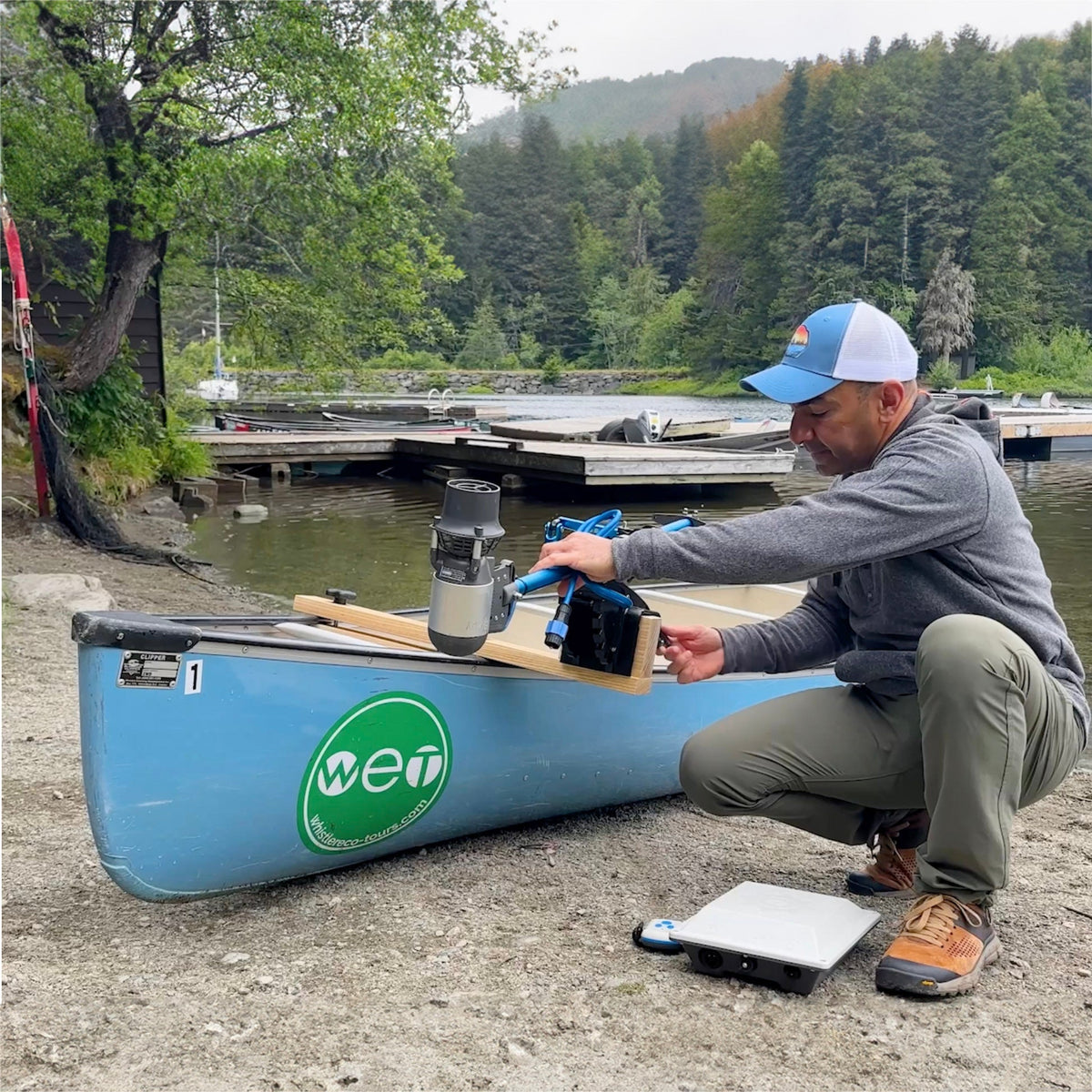 Person setting up equipment on a canoe by a lake with trees and mountains in the background