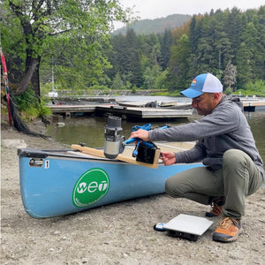 Person setting up equipment on a canoe by a lake with trees and mountains in the background