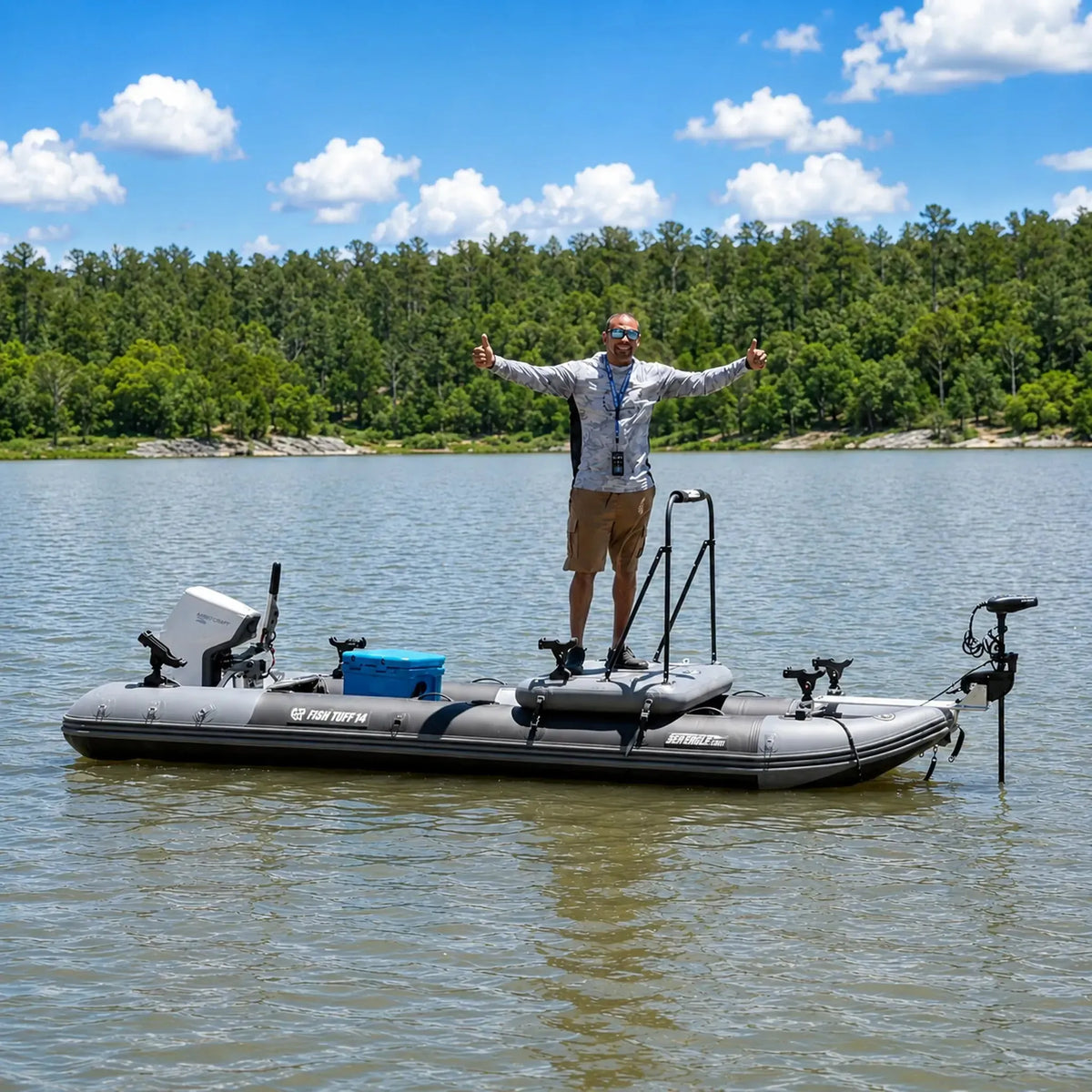 A man stands on a gray inflatable fishing boat with a motor, photographed on open water with a forest shoreline and blue sky in the background.