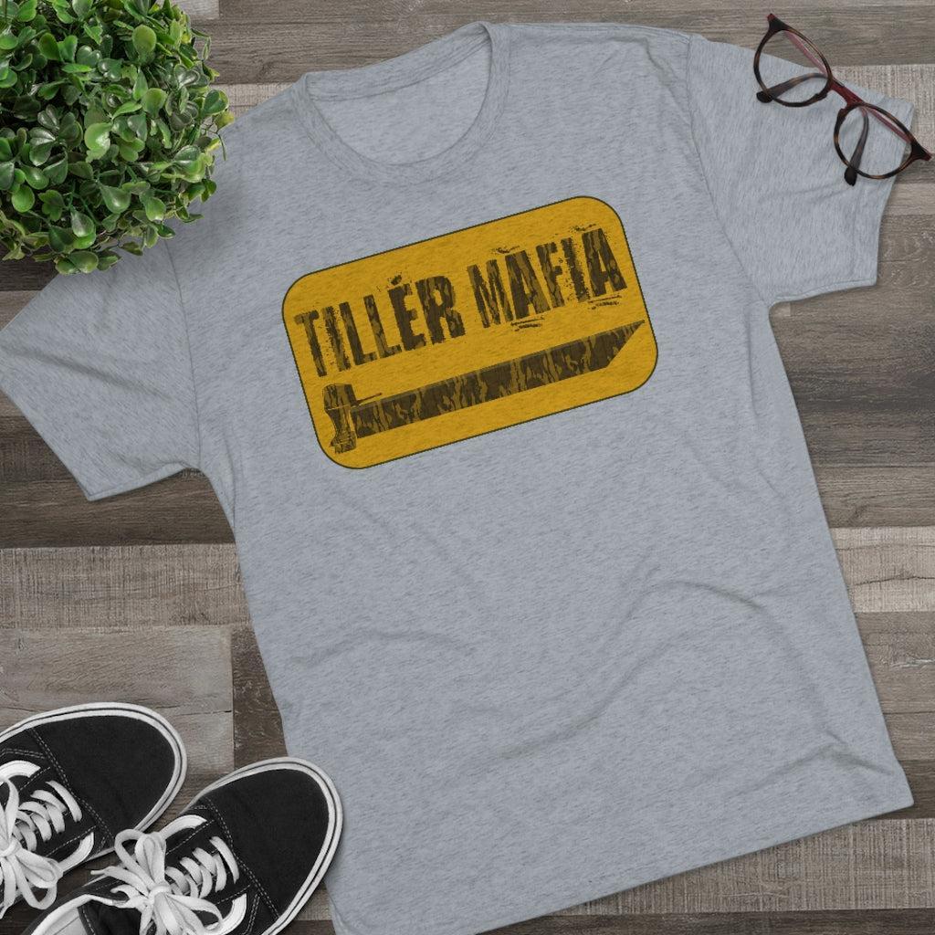 A heather gray t-shirt with a yellow and black distressed graphic reading 'TILLER MAFIA' laid flat on a wood floor with decorative props including a plant, glasses, and black sneakers.