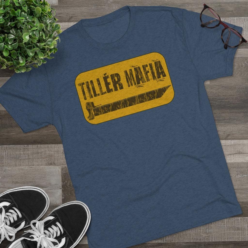 A heathered blue crewneck t-shirt with a yellow road-sign graphic reading 'TILLER MAFIA' and a wrench, styled in a flat lay on a wood floor with a plant, glasses, and black sneakers.