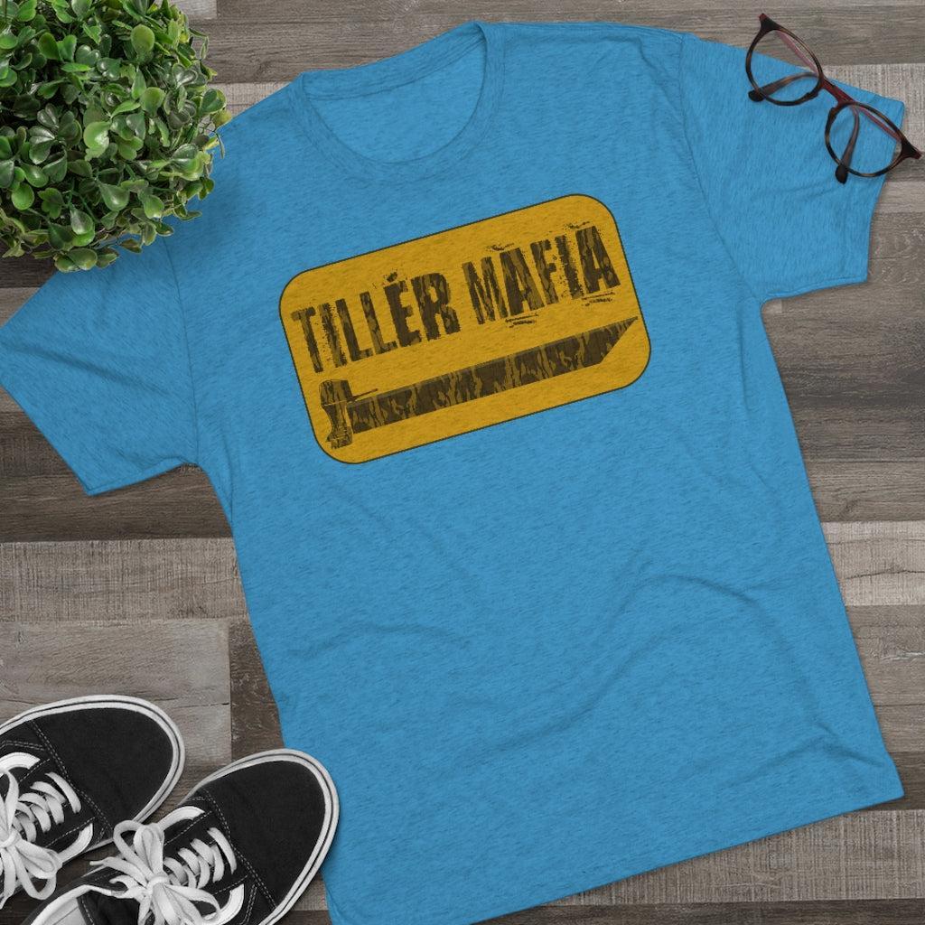 A heather blue graphic t-shirt laid flat on a wooden surface, featuring a yellow sign-style graphic that reads 'TILLER MAFIA' with a pencil illustration. Black sneakers, eyeglasses, and a green plant are styled around the shirt.