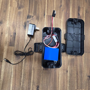 Max It Out LEDs Max Kayak Power System open battery enclosure displaying the internal blue battery pack, wiring harness, power lead, and an AC wall charger on a wooden surface.