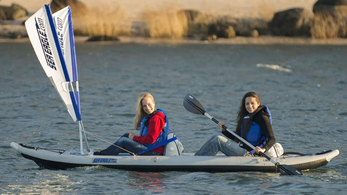 Two women in a gray Sea Eagle FastTrack inflatable kayak with a white and blue sail, paddling on calm water.