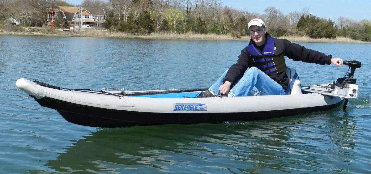 Man in a gray and black Sea Eagle 385ft FastTrack™ inflatable kayak with a motor, on calm water.
