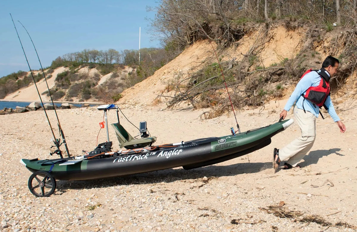 Man pulling a dark green Sea Eagle 385fta FastTrack Angler inflatable kayak with fishing rods on a sandy beach.