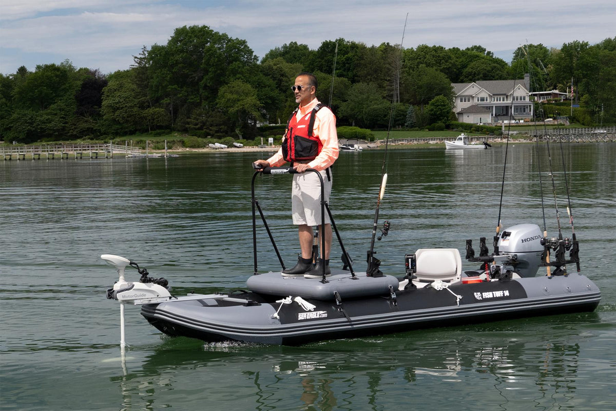 A man stands on a gray sea eagle inflatable fishing boat with a mounted motor, fishing rods, and a white outboard motor, on the water with a shoreline, trees, and houses in the background.