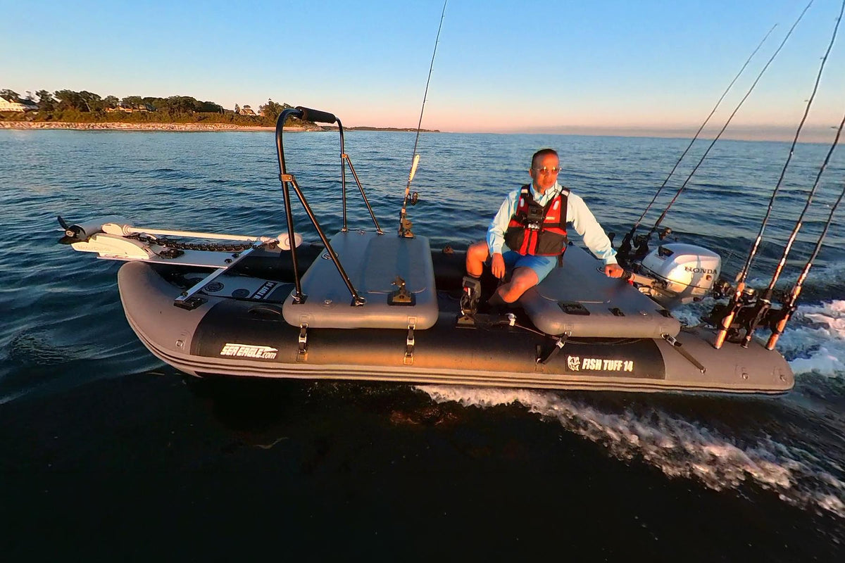 A man wearing a life jacket sits on a gray sea eagle inflatable fishing boat with multiple fishing rods mounted on the sides, moving across open water during sunset.