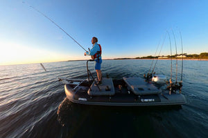 A man stands on a sea eagle fishing boat on open water at sunset, holding a fishing rod. The boat is equipped with multiple fishing rods and tackle gear, and the water reflects the blue sky and warm light.