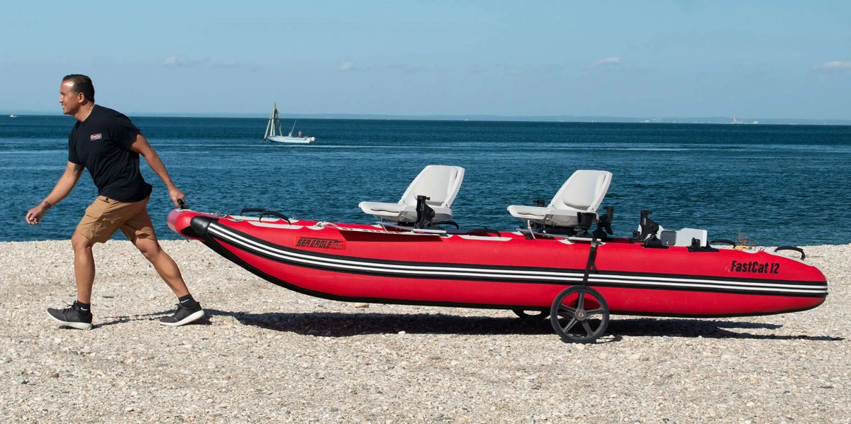 Man pulling a red Sea Eagle FastCat12 inflatable catamaran with two white seats on a pebbly beach.