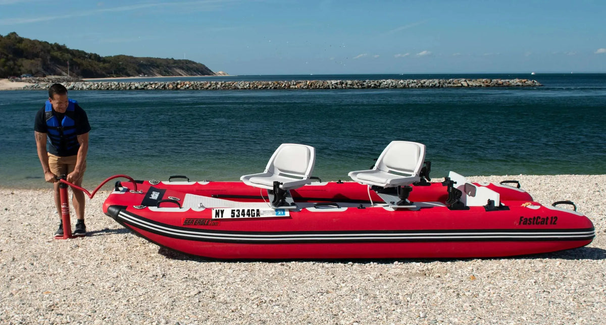 Red Sea Eagle FastCat12 inflatable catamaran boat with two white seats on a gravel beach, being inflated by a man.