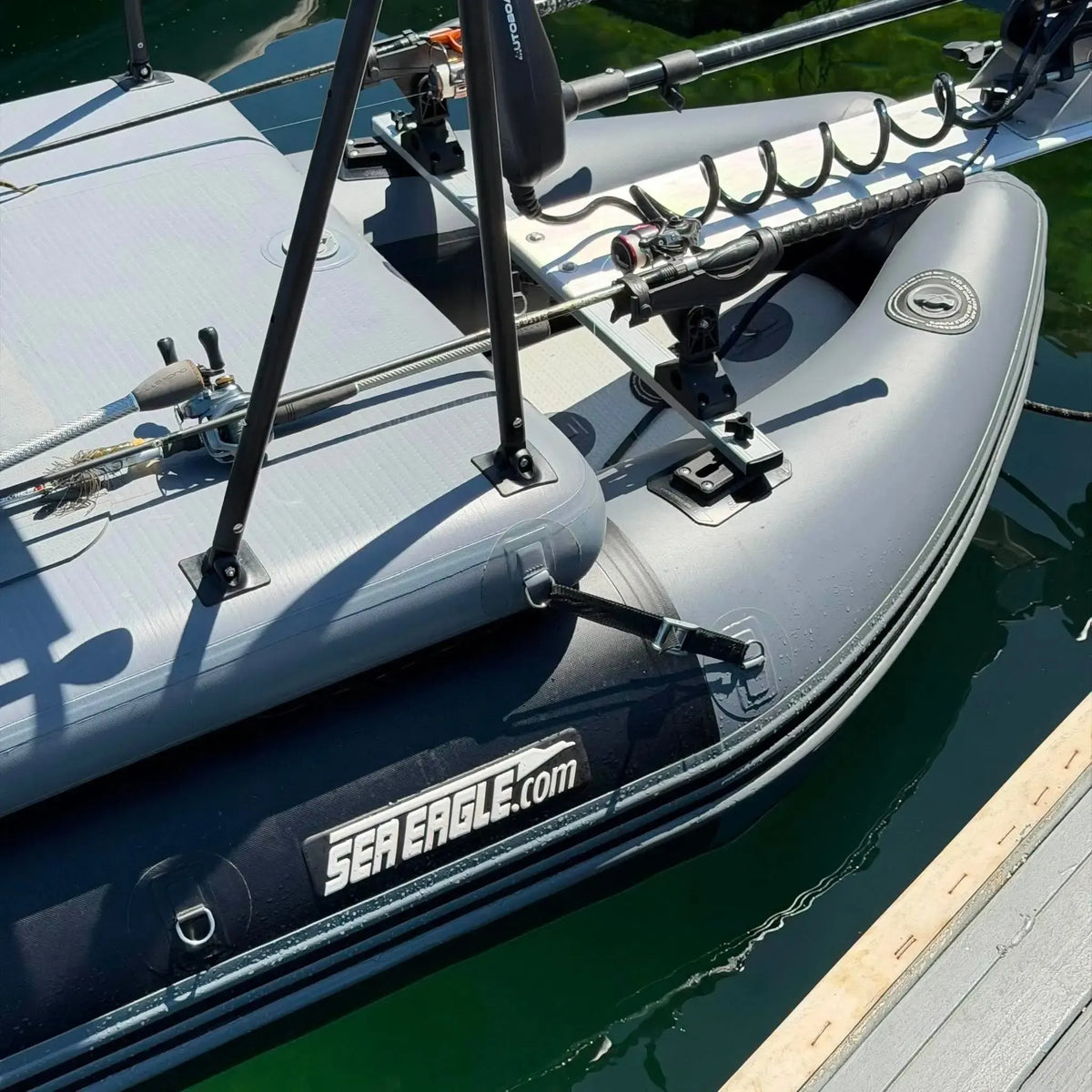Close-up of a gray inflatable boat docked at a pier, showing the side branding 'SEA EAGLE.com' and fishing rod holders mounted along the deck.