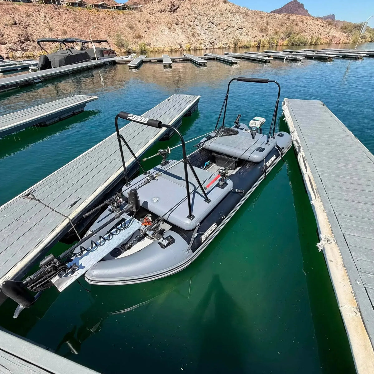 A gray sea eagle inflatable boat with a black frame and trolling motor attachment, docked at a wooden marina on calm greenish-blue water with rocky mountains in the background.