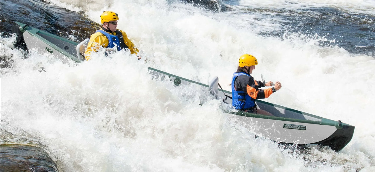 Two people in yellow helmets and life vests paddling a grey and green Sea Eagle TC16 inflatable canoe through whitewater rapids.