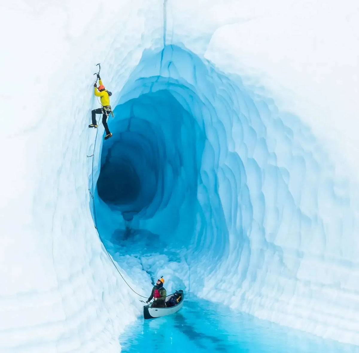 Two adventurers in a bright blue ice cave, one climbing the icy wall, the other in a white inflatable canoe.
